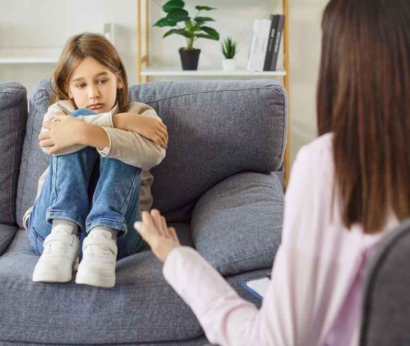 Child sitting with arms crossed on couch as therapist answers common ABA therapy questions at home