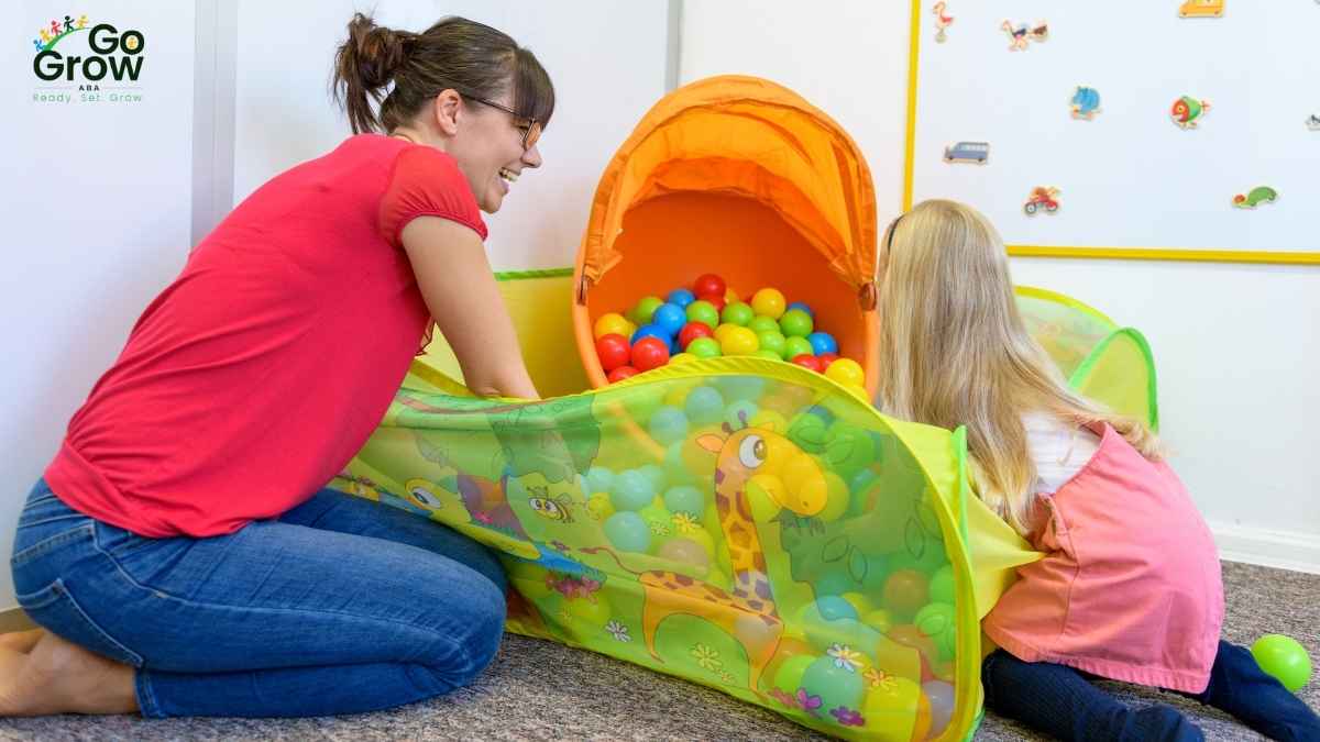 Therapist smiling while playing with a young child at a colorful ball pit on the floor